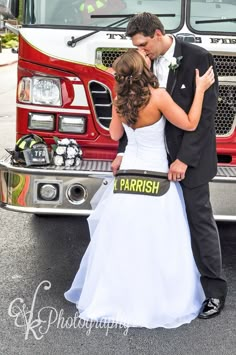 a bride and groom standing in front of a fire truck with their arms around each other