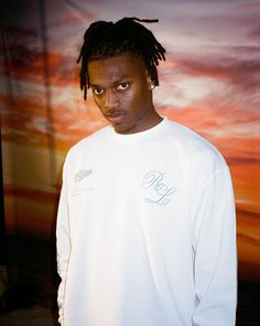 a young man with dreadlocks standing in front of a sunset background wearing a white long sleeved shirt