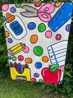 a person holding up a painting with school supplies on it in front of some bushes