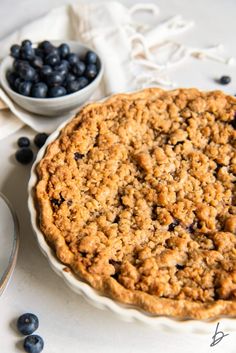 a blueberry crumb pie sitting on top of a table next to a bowl of blueberries