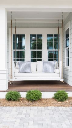 a porch swing with pillows on it in front of a white house that has large windows