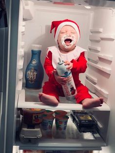 a baby dressed as santa claus sitting in an open refrigerator, holding a bottle of milk