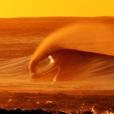 a large wave in the middle of an ocean at sunset, with orange and yellow hues