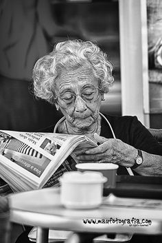 an old woman reading a newspaper while sitting at a table with a cup of coffee