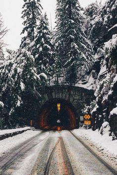 a snow covered road going into a tunnel with trees on both sides and light at the end