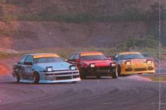 three cars driving on a race track with hills in the backgrouds behind them