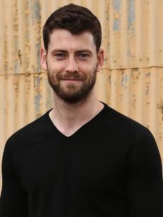 a man with a beard standing in front of a rusty wall wearing a black v - neck shirt