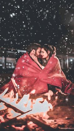 a couple wrapped in a blanket sitting next to a campfire on a snowy night