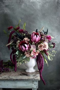 a white vase filled with lots of flowers on top of a wooden table next to a gray wall