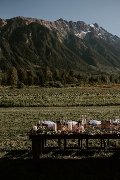 an outdoor table set up in the middle of a field with mountains in the background