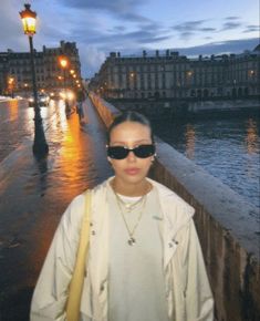 a woman wearing sunglasses standing on a bridge next to the water at night with buildings in the background