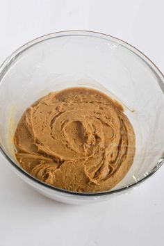 A glass bowl containing smooth, light brown gingerbread dough wrapped with plastic on top, set against a plain white background.