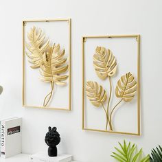 two gold leaf wall art hanging on the wall next to a black vase and potted plant