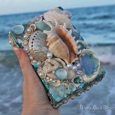 a hand holding up a small box with shells and pearls on the beach next to the ocean