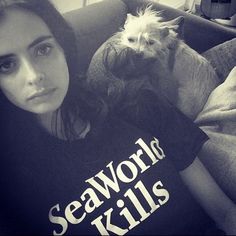 a woman sitting on a couch holding a small white dog in her lap and wearing a t - shirt that says seaworld kills