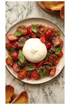 a white plate topped with tomatoes and basil next to slices of bread on top of a marble counter