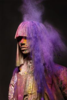 a woman with purple hair and makeup is covered in colored powder as she stands against a black background