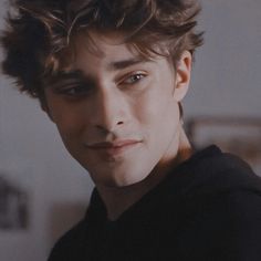 a young man with curly hair and blue eyes looks into the camera while wearing a black shirt