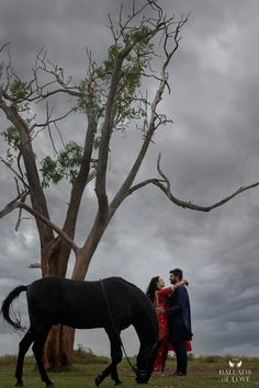 a man and woman standing next to a tree with a horse in the foreground