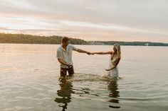 a man and woman holding hands in the water with trees in the background at sunset