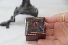 a hand holding a small wooden box with an eye on it's side and a statue in the background