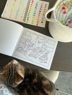 a cat laying on top of a table next to an open book and crayons