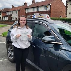 a woman standing next to a blue car holding up a check for her driving license