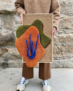 a person holding up a piece of artwork made out of grass and blue flowers in front of a stone wall