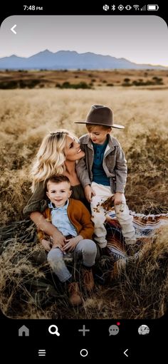 a woman and two children are sitting on the ground in a field with mountains behind them