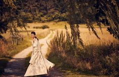 a woman in a white dress walking down a dirt road next to tall grass and trees