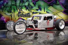 an old fashioned hot rod car is parked in front of a wall with graffiti on it