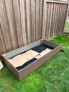 an empty box sitting in the grass next to a wooden fence with some cardboard on it