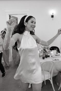 a woman in a white dress is dancing with her hands up and people are standing around the table