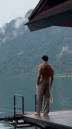 a shirtless man standing on a dock looking out at the water and mountains in the distance