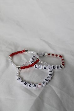 two personalized bracelets with red beads and white letters are on a bed sheet