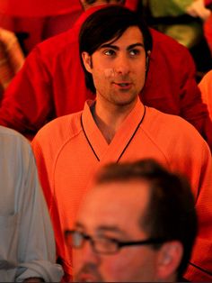 a man in an orange shirt sitting next to another man with glasses on his head