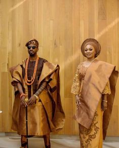 two people standing next to each other in front of a wooden paneled wall wearing african clothing