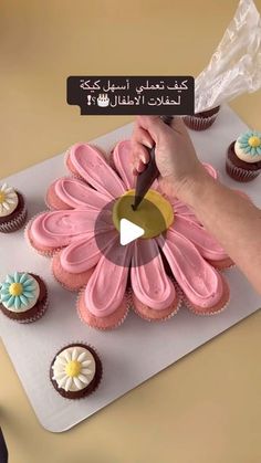 a person cutting a pink flower shaped cupcake on top of a white board with other cupcakes around it