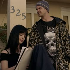 a man standing next to a woman with a book in her hand and a skull t - shirt on