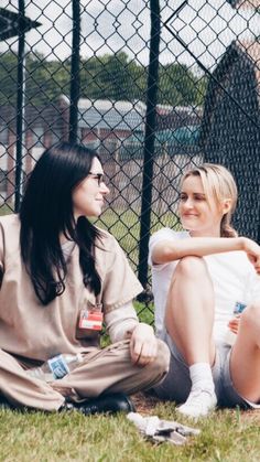 two women sitting next to each other on the ground in front of a chain link fence