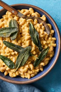 a blue bowl filled with macaroni and cheese topped with sage leaves next to a wooden spoon