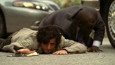 a man laying on the ground next to a car and another man looking at his cell phone