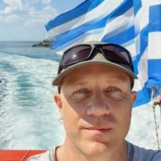 a man wearing sunglasses and a hat on a boat in the ocean with a greek flag behind him