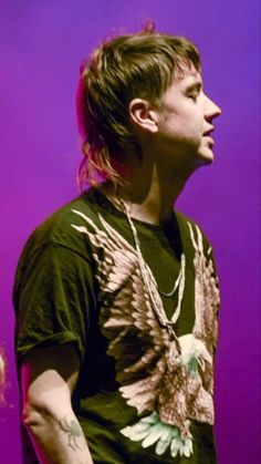 a young man with long hair and an eagle t - shirt standing in front of a purple background