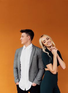 a man and woman standing next to each other in front of an orange wall with their hands on their chins