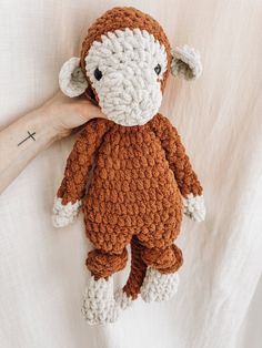 a hand holding a brown and white crocheted monkey stuffed animal on top of a bed