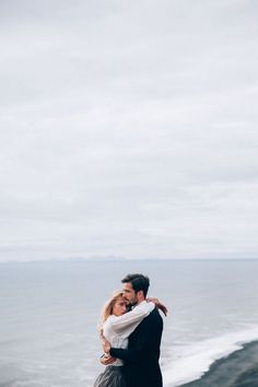 a man and woman standing on top of a cliff next to the ocean holding each other