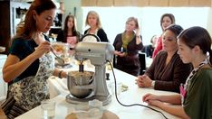 a group of women standing around a kitchen counter with a mixer on it's stand