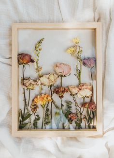 an arrangement of dried flowers in a wooden frame on a white bed linens background