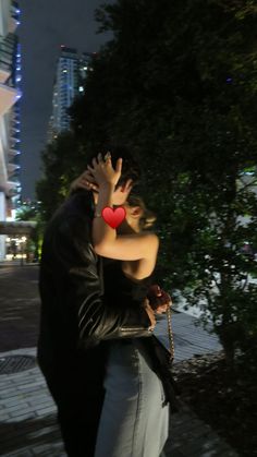 a man and woman embracing each other in front of a building at night with the city lights lit up behind them